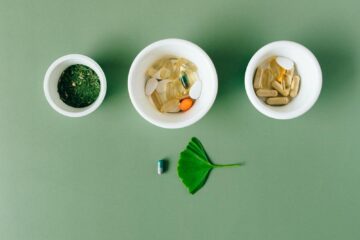 Flatlay of capsules and herbs in ceramic bowls on green surface.